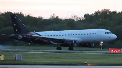 A plane with a white body and blue tail with white stars is seen from the side on a runway