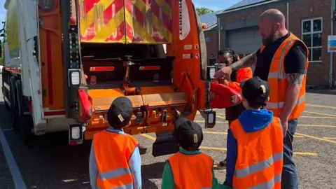 Contributed Three young boys face away from the camera looking at the rear of a bin lorry. A man is standing next to them, speaking with them and pointing at part of the lorry. They wear matching black caps and orange hi-vis jackets. 