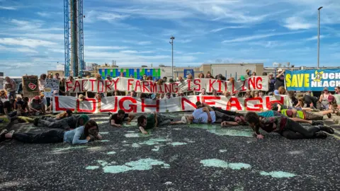 Slí Eile People lie on the ground next to mud puddles, while in the background people hold up a large banner with the inscription "Factory farming is killing Lough Neagh".