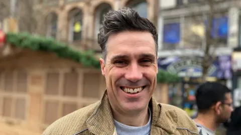 Darren is smiling and is standing in front of the Christmas Market in Leicester. He has black hair and is wearing a brown-ish checked jacket. 