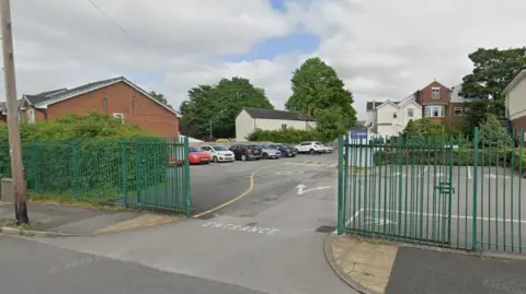 Google A Google street view image of the entrance to a school car park. Green fences are situated either side of the entrance. Several buildings can be seen in the background alongside a line of parked cars.