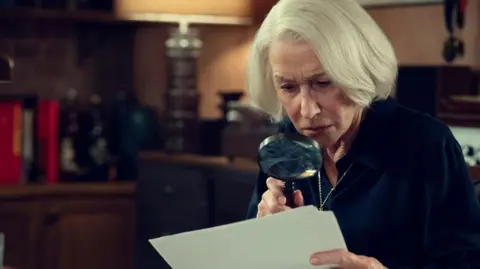Helen Mirren is inspecting a document with a magnifying glass whail sat on a brown leather arm chair