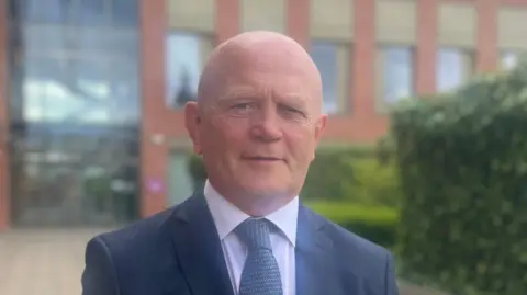 A bald man in a suit standing outside in front of a large red brick building with several windows. It is daytime and some hedges can be seen to his right.