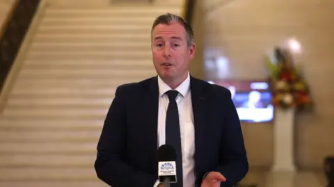 PA Media a man in a black suit blazer with a skinny black tie over a white shirt, speaking at a Northern Ireland Assembly - branded microphone in a room that is recognisable as Stormont's great hall