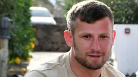 Scot, a man with short brown hair and stubble looking into the camera. He's wearing a beige top and stood in a garden.  