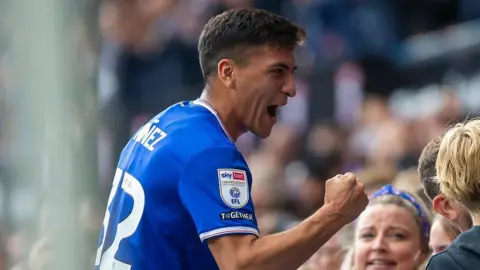Getty Images Marcelino Nunez wearing a blue and white Ipswich Town strip celebrates with fans in the stands after a goal. He has one fist slightly raised in front of him as he celebrates. 