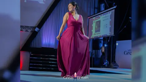 Miss Great Britain/Jin Rathood Elle Seline in a long pink dress onstage. She is posing, holding the dress out at each side. There is a projector screen behind her with her name on it and a photo.