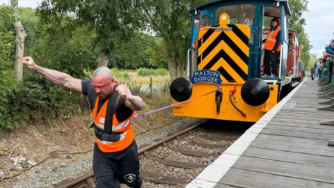 The strongman is at the finish line on the tracks leaning forward with both arms up in the air.  The locomotive is following behind via a rope.