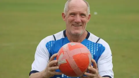 Roy holding a red football wearing a blue and white Posh jersey