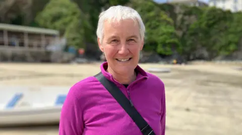 Barbara Mortimer has short grey hair and wearing a purple top. She is standing outside on a beach