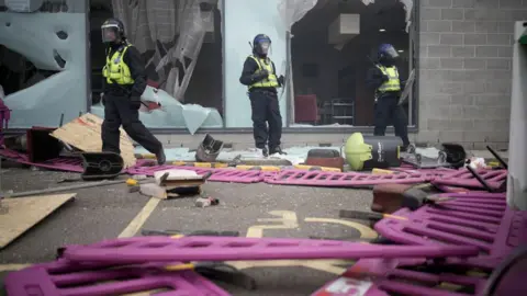 PA Media Three police officers in riot clothing stand next to smashed windows at the hotel. Several barriers are on the ground around them along with other items thrown towards them.