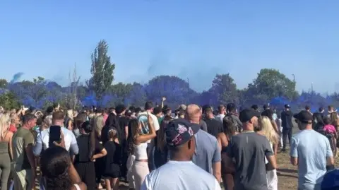 A crowd of young people can be seen standing in a field in the late afternoon sunshine. There is blue smoke above their heads and trees can be seen in the distance.