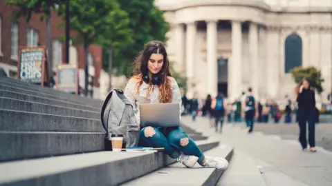 Getty Images A miss  wearing bluish  jeans and a stripy achromatic  and achromatic  garment  is sitting connected  factual  steps connected  her laptop. She has a container  and a java  beside her. She is extracurricular  an aged  grey gathering  with radical   walking past. 