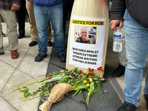 A poster reading 'justice for rob who died whilst under extreme stress waiting for his licence'