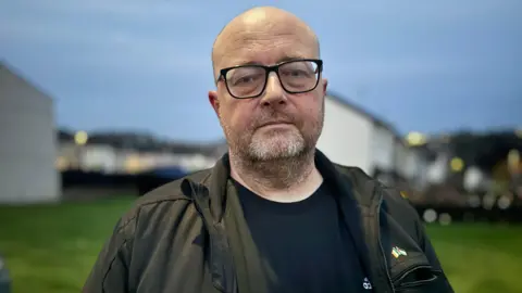 Bald man wearing glasses and wearing green jacket looking straight ahead as his picture is taken