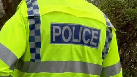 BBC A police officer wearing a yellow jacket with a sign reading police on the back walking away from the camera