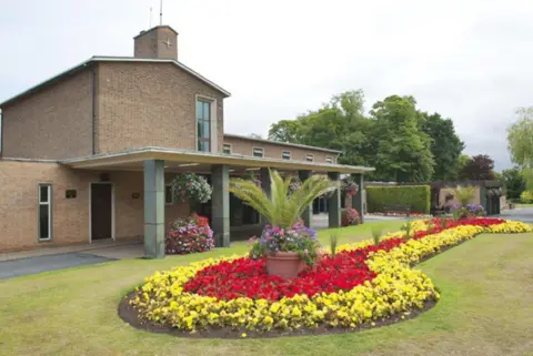 City of Wolverhampton Council The crematorium building is on the left and in the centre of the photo. Flowers are in the foreground in front of the building and trees are on the right of the image in the background.