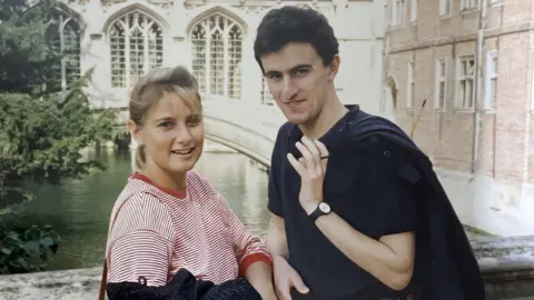Rose Grant with her boyfriend Tim Burman. She is wearing a red and white stripey long-sleeved top. Tim is wearing a dark polo shirt and is holding a jacket over his left shoulder. They are stood on a bridge over a river or canal with old buildings behind them.