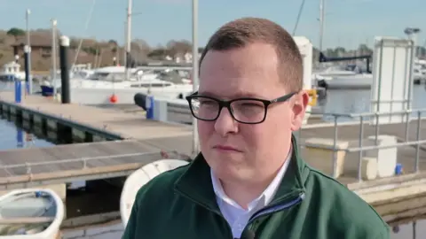 Jack is standing by the water at Lymington Quay. He is wearing a white shirt with a green jacket. There are small boats and masts in the background.