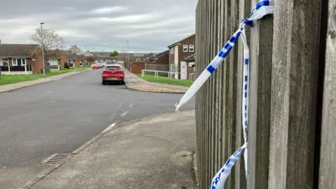 A residential street with a fence with blue and white police tape tied around it on the right hand side. Homes on either side of the street and cars parked along the street in the distance.