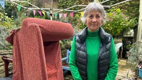 Ms Rees stands in a paved area of her back garden, wearing a green jumper and black gilet, looking into the camera. Next to her, a red sofa is tipped on one end, drying out.