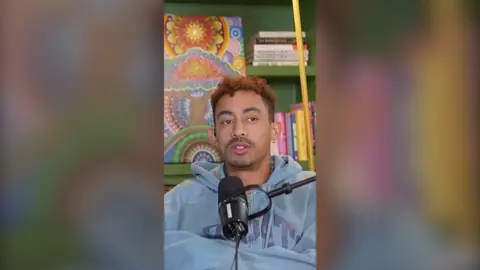 Jordan Stephens is wearing a light blue hoodie and sitting behind a microphone. He has short, curly brown hair.