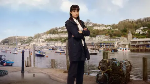 Red Planet Pictures Zahra Ahmadi as the character Det Sgt Esther Williams posing for a press shot with Looe behind her across the water on which yachts are moored. She has long dark hair and is wearing a navy anorak and trousers with a white shirt. She is smiling and has her arms crossed. There are lobster pots at her feet.