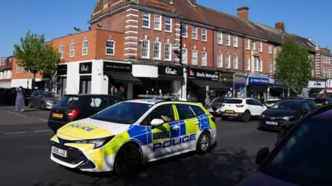 A police car parked on a street surrounded by other cars and in front of a row of shops 