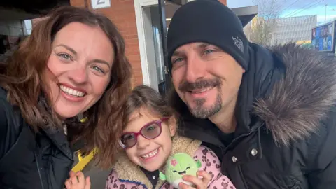 Family handout Three people all together smiling and looking at the camera. The young girl in the middle is holding a toy and has on a pink and brown leopard print coat, with purple glasses on. A woman is to the left, with long dark hair and a man to the right with a black hat on, and black coat with a furry hood. 