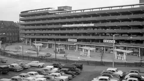 A black and white image of a concrete multi-storey car park with Tesco signs on the ground floor and a petrol stion fore court