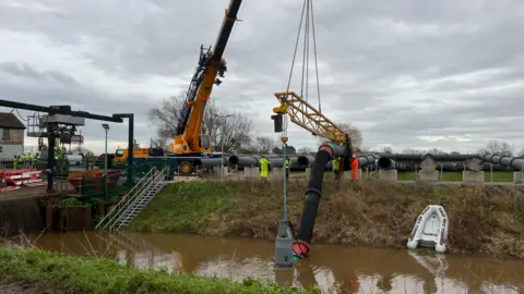A row of large pipes above a river. One pipe is being connected to the river via a large hose. An industrial facility with two cranes is visible behind.