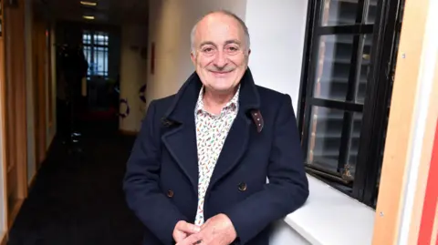 Tony Robinson stands in a corridor wearing a navy coat over a white shirt with birds on it. 