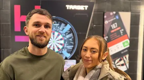 Adam Kenyon (left) with short brown hair and beard wearing an olive top and Jade Pettiner with long brown hair and wearing a pale pink and grey scarf and beige coat stand in front of a dart board. They are smiling.