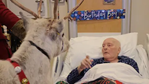 St Helena Hospice Mick Hodgson lies in a hospital bed in blue pyjamas, he is looking at a reindeer which has bells around its neck. 