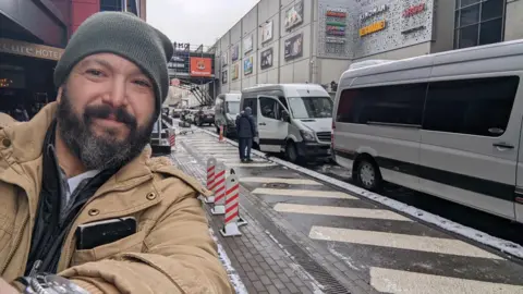 Grey Bull Rescue/Handout Bryan Stern, a man with a brown and grey beard wearing a khaki wool hat and tan-coloured jacket, takes a selfie in front of three vans and a shopping centre