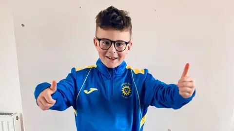 Justine Moseley Leo Moseley stood with his thumbs up looking at the camera. He is wearing glasses and is smiling. He is wearing a blue and yellow tracksuit top.
