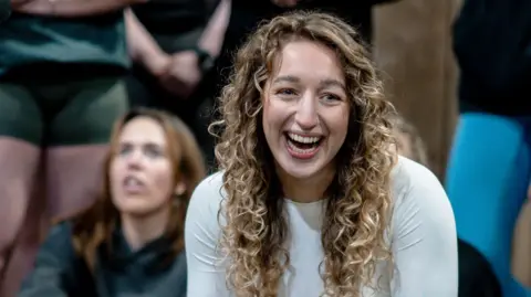 Craig Williams Sydney has long blonde wavy hair and is wearing a white long-sleeved gym t-shirt. She is in a busy studio with unidentified females behind her.