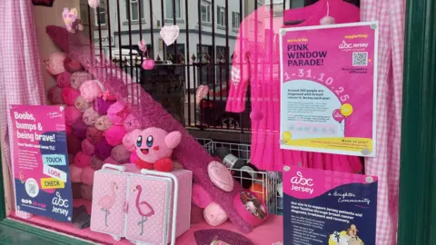 A window display of a shop in Jersey with three posters about breast cancer awareness, with different colours of pink yarn on top of each other, a pink shirt hanging up and the pink kirby toy on top of a pink flamingo sewing box.