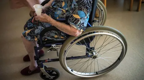 AFP via Getty Images An elderly lady holding a plastic water cup sits in a wheelchair in a care home