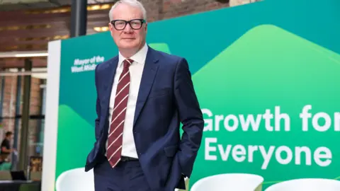 A man wearing a navy suit and tie stands in front of a large green billboard. He has his hands in his pockets. The words "Growth for... Everyone" can be seen in big white letters.