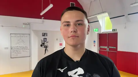 Nicholas stares straight into the camera, wearing a black t-shirt and standing in his gym where he trains in Hull.