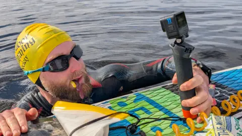 Calum Maclean Wild swimmer Calum Maclean in the water with just his head and shoulders visible leaning on a paddle board. He is wearing a yellow swim cap and black goggles and a black wetsuit with red and blue design. He is holding a gopro camera. Image was taken around Inchyra in September this year.