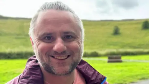 A man wearing a dark red rain coat is standing outdoors with a green field behind him. He has grey hair and a stubbly beard. He is smiling for the photograph.