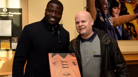 Ed Bagnall Two men in the reception of Wolves football club. Both wearing coats. One has a copy of a book about football shirts