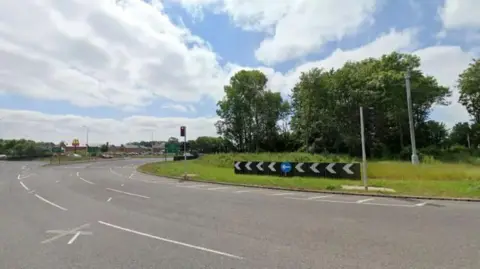 Google Streetview image of a three-lane roundabout, which has trees and grass in the middle.