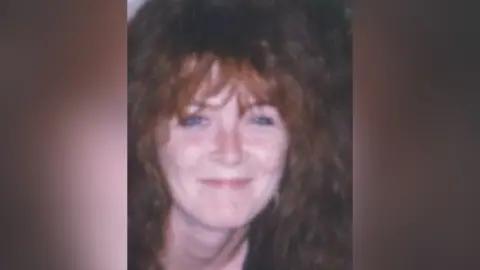 A blurry image of a woman with long, wavy red hair. She is smiling at the camera. The background of the image is blurred.