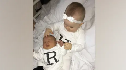 Family handout Harper, a young toddler, sits on a hospital bed holding newborn baby Romie. They are both wearing white baby grows with their initials on the front in black lettering. 