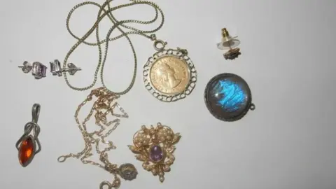 Photo of several items of jewellery taken on a white table. The items include a gold necklace with a gold coin charm. Other items include a pearl broach with purple stones in the centre.