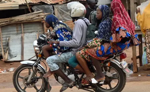 Anadolu/Getty Images Quatro meninas em roupas estampadas coloridas com lenços de cabeça em uma moto dirigida por um jovem em uma camiseta cinza e capacete branco visto recentemente em uma rua em Lomé, Togo.