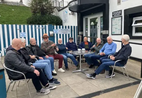 Ten men sit outside a cafe on metal chairs, they are talking and drinking cups of tea.